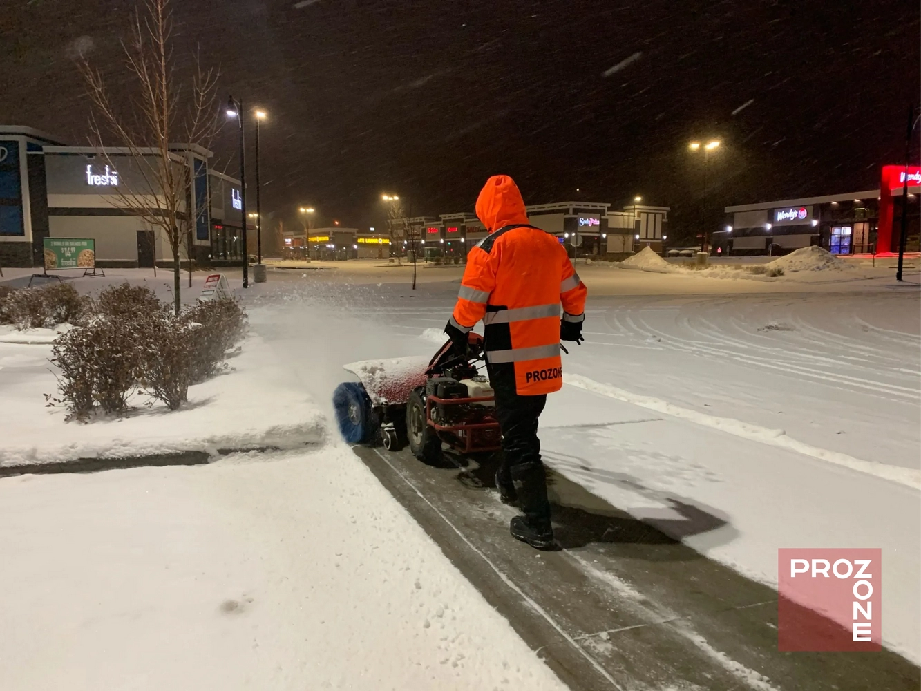 Worker operating sidewalk machine during snow removal near shopping center, supporting parking lot snow removal with essential winter supplies.