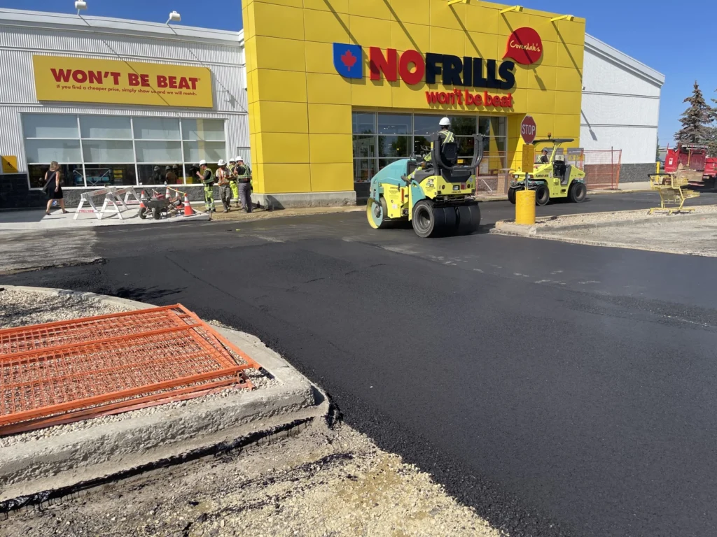 Construction crew completing asphalt repair on a parking lot area in front of a yellow retail building.