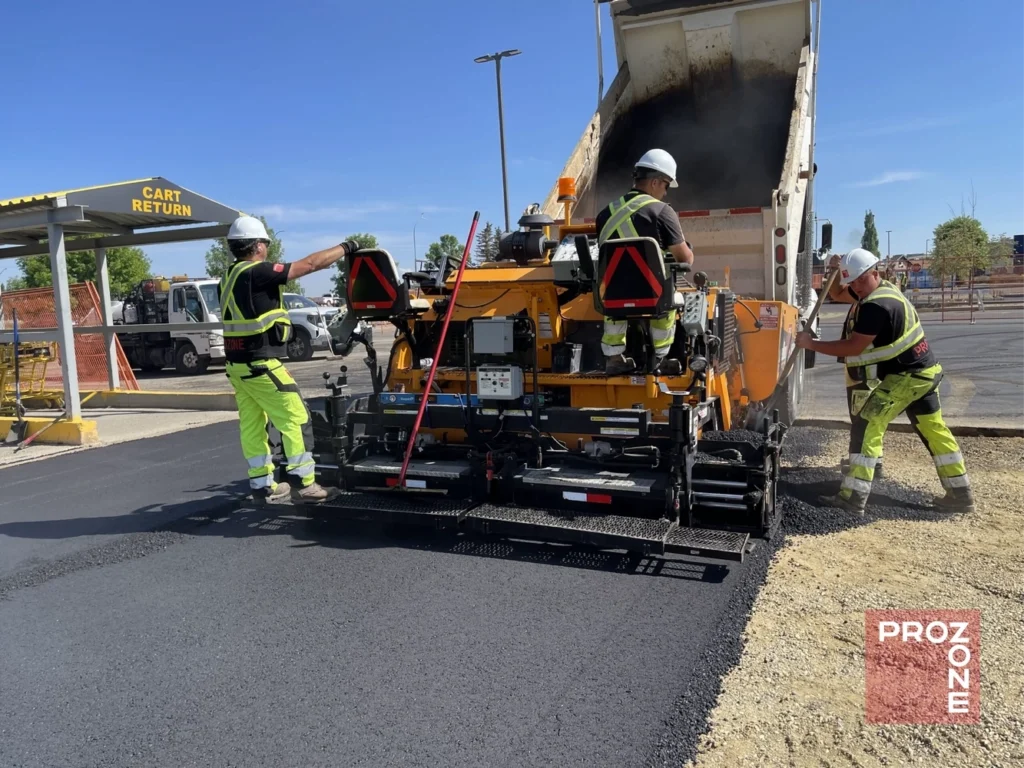 Crew members operating heavy equipment spreading asphalt mix during detailed asphalt repair on a parking lot.